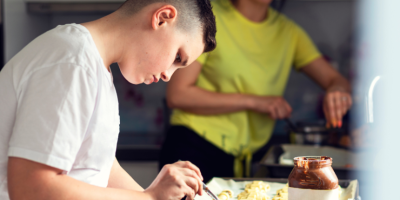 Young boy is cooking in a kitchen, watched over in the background by a female adult